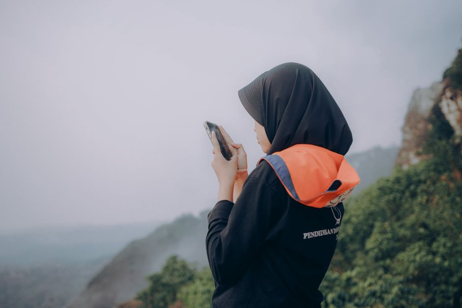 A woman in a hijab stands outdoors, using a smartphone in the Indonesian mountains.