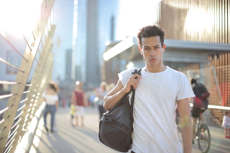 Cool confident African American male in earbuds and casual clothes looking at camera with interest while standing on street with large black bag on shoulder and listening to music against blurred people and urban environment in city