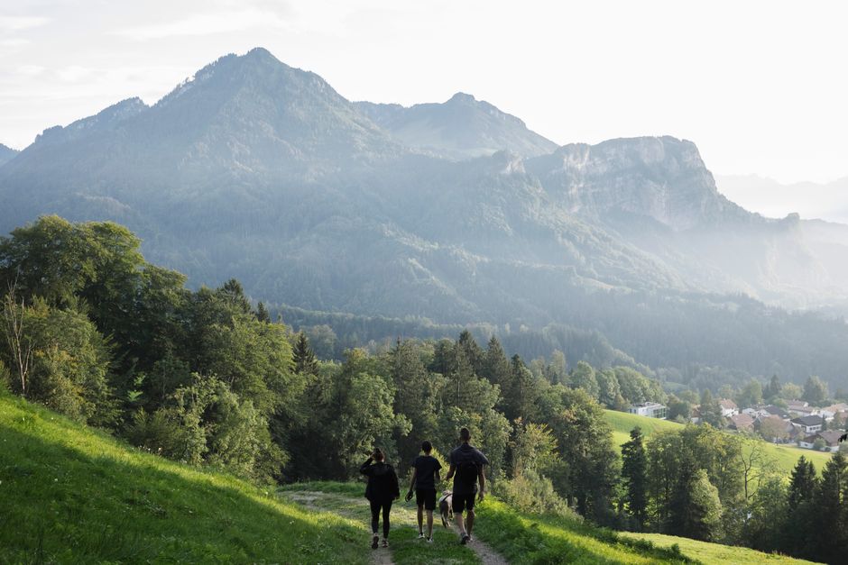 Three people hiking on a trail with scenic mountain view, lush greenery, and distant village.