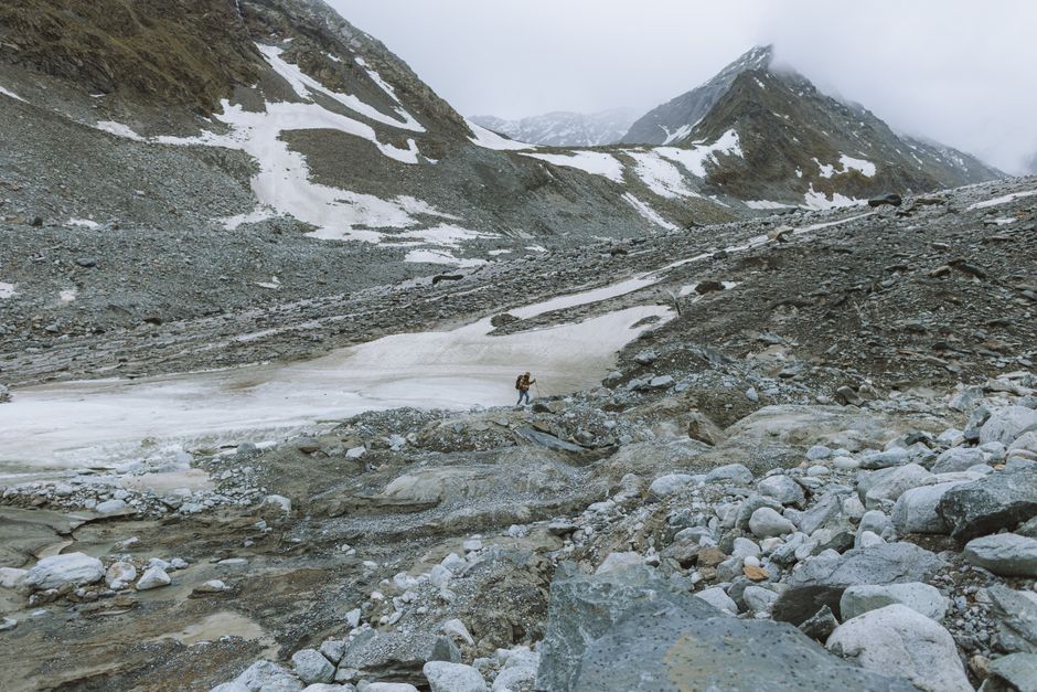 A lone hiker explores a rocky, snow-dusted mountain landscape under cloudy skies.
