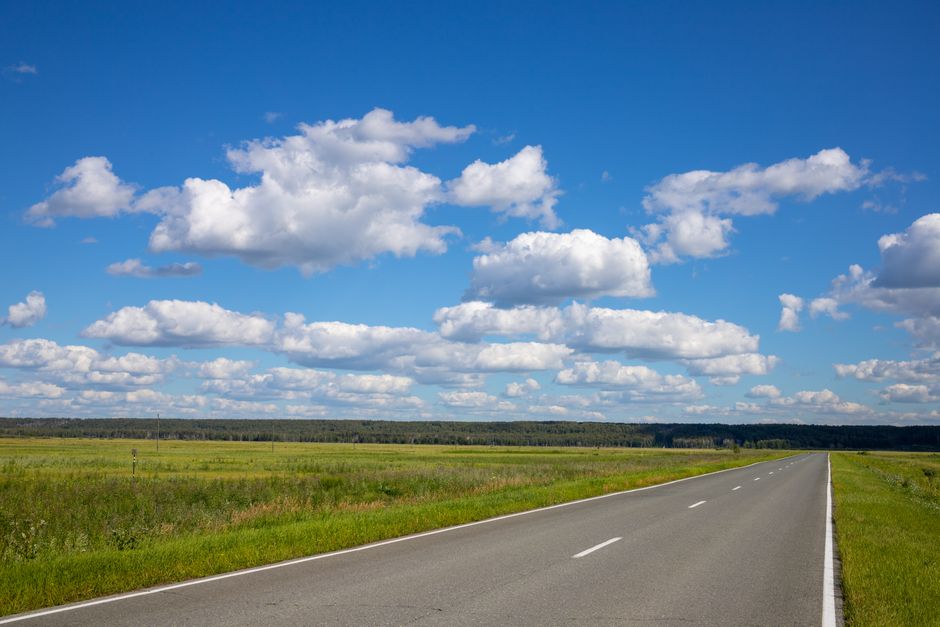 A scenic country road extends through a sunny rural landscape under a sky filled with fluffy clouds.