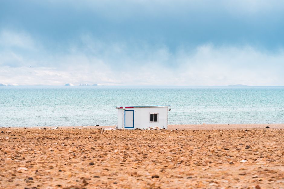 A lone white hut stands on a serene beach with the vast blue ocean in the background.